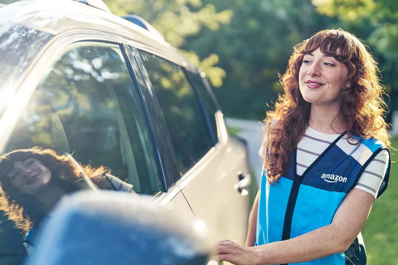 A young woman stands next to a silver vehicle and has on hand on the driver's side door handle.