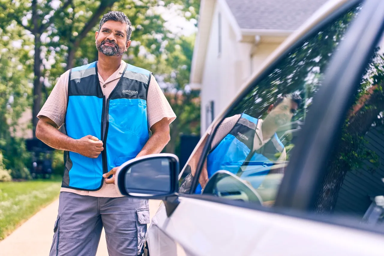 A man stands next to a vehicle and zips up a blue Amazon vest.