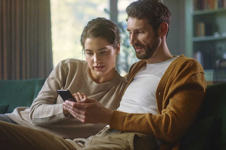 A couple sits on a living room couch and look down at a phone in the man's hands
