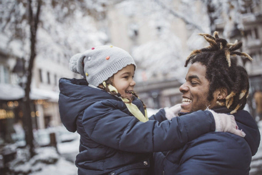 A man holds a child up and they laugh together. They are outside in a snow-covered neighborhood and they wear winter gear.