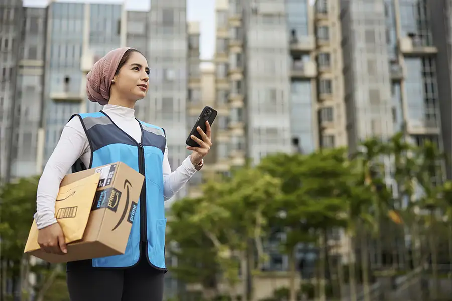 A woman in a blue Amazon vest stands near a series of condo buildings. She holds a phone in on one hand and two packages in the other.
