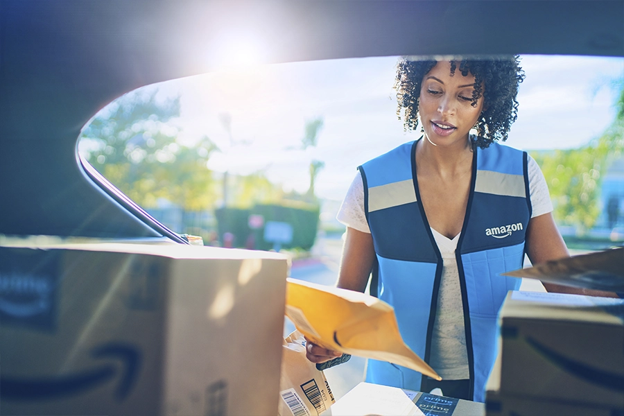 A woman stands behind the open trunk of her SUV and looks down at a package. She wears a blue Amazon vest, and in the back of the trunk are half a dozen more packages.