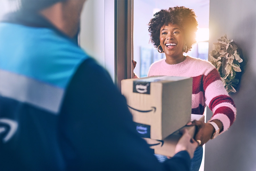 A man in a blue Amazon vest hands a woman two packages. She smiles and stands in front of an open front door.