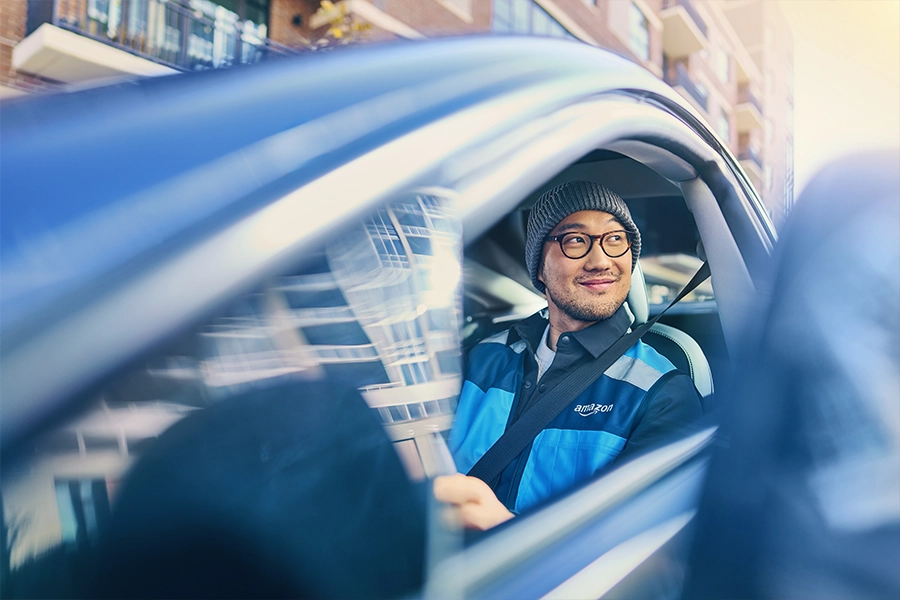 A man in a blue Amazon Flex jacket sits in the drivers seat of a vehicle and looks out the window