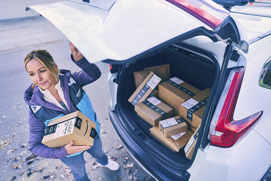 A woman in a blue and purple Amazon Flex jacket stands behind her car with the hatch open. In the back of the car are around a dozen packages and the woman carries one package.