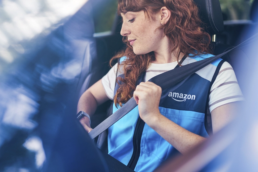 A young woman who wears a blue Amazon Flex vest sits in the driver's seat of a car and buckles up her seat belt.