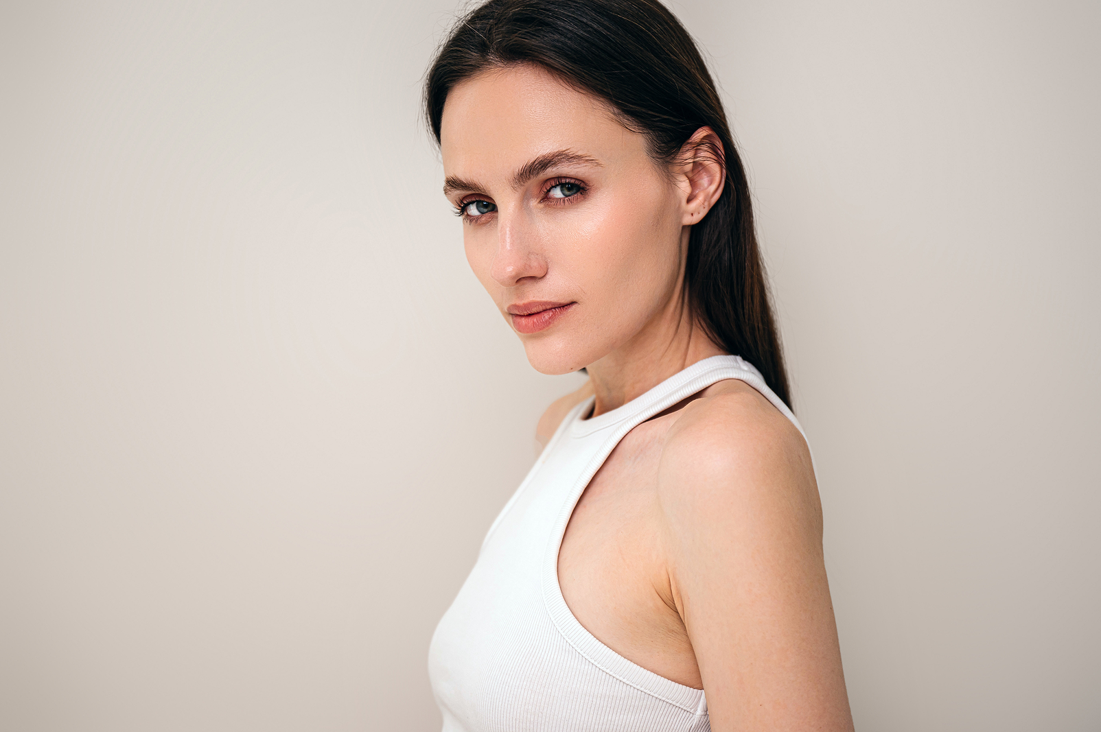 Portrait of a woman with long dark hair wearing a white sleeveless top against a plain beige background.