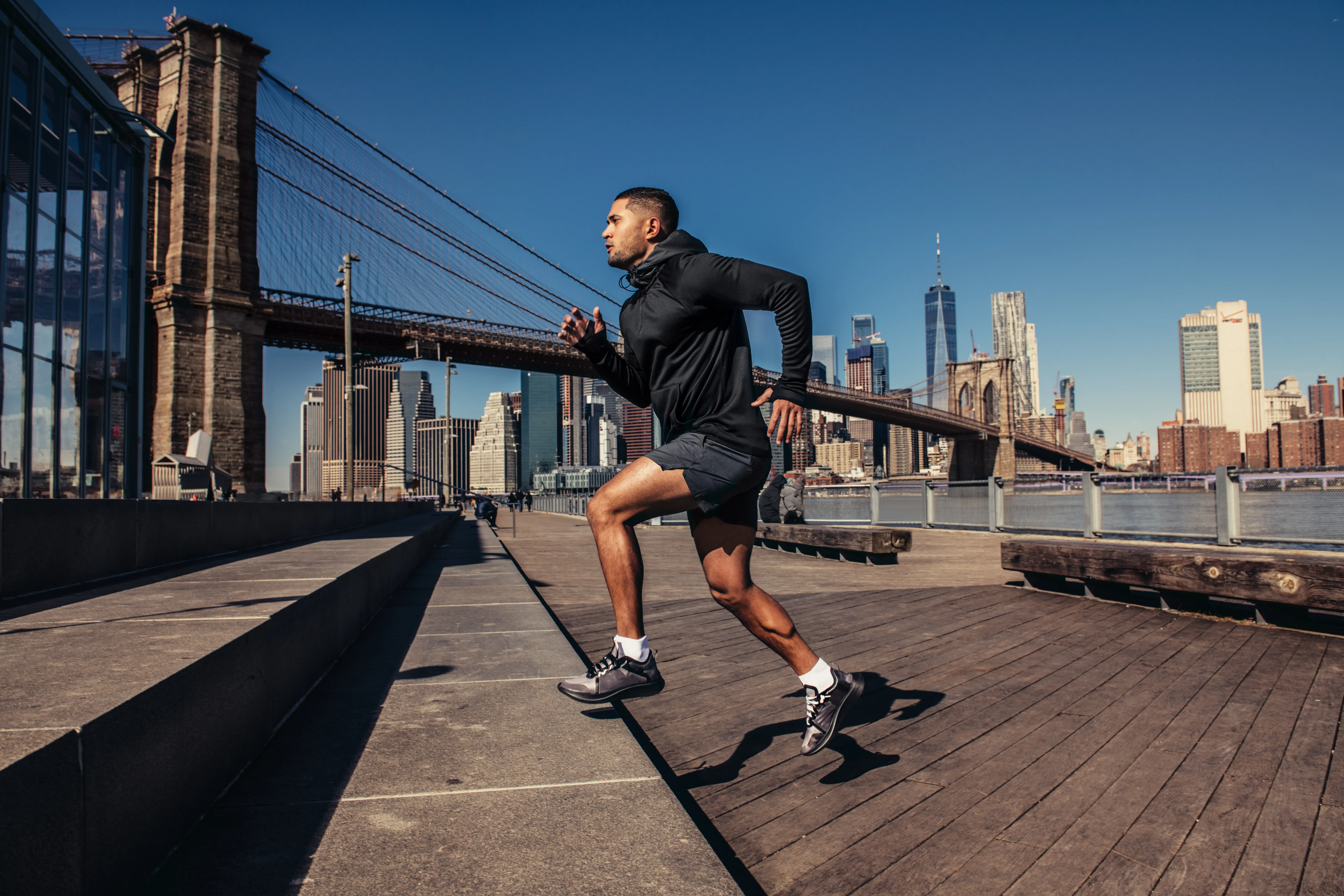 Athletic man running outdoors near Brooklyn Bridge with New York City skyline in the background on a clear day.