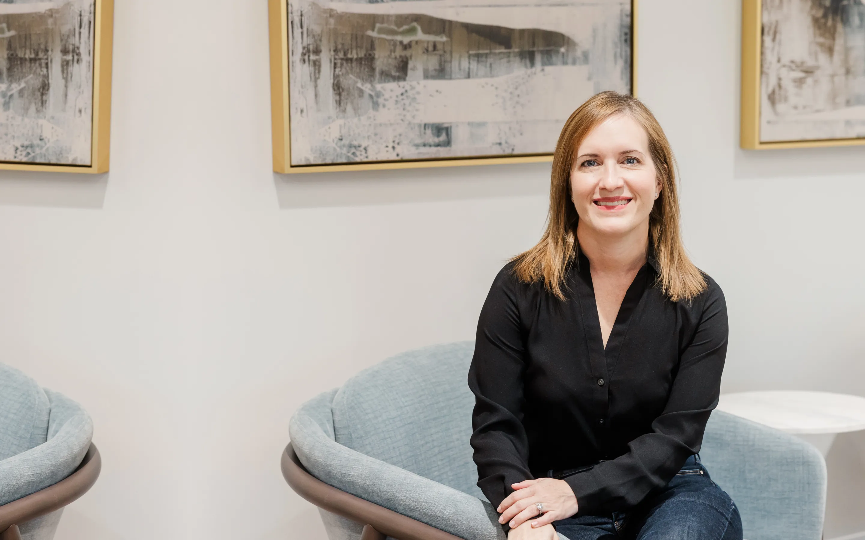 Smiling woman with blonde hair in a black shirt sitting on a light blue chair in a bright room with abstract paintings on the wall.