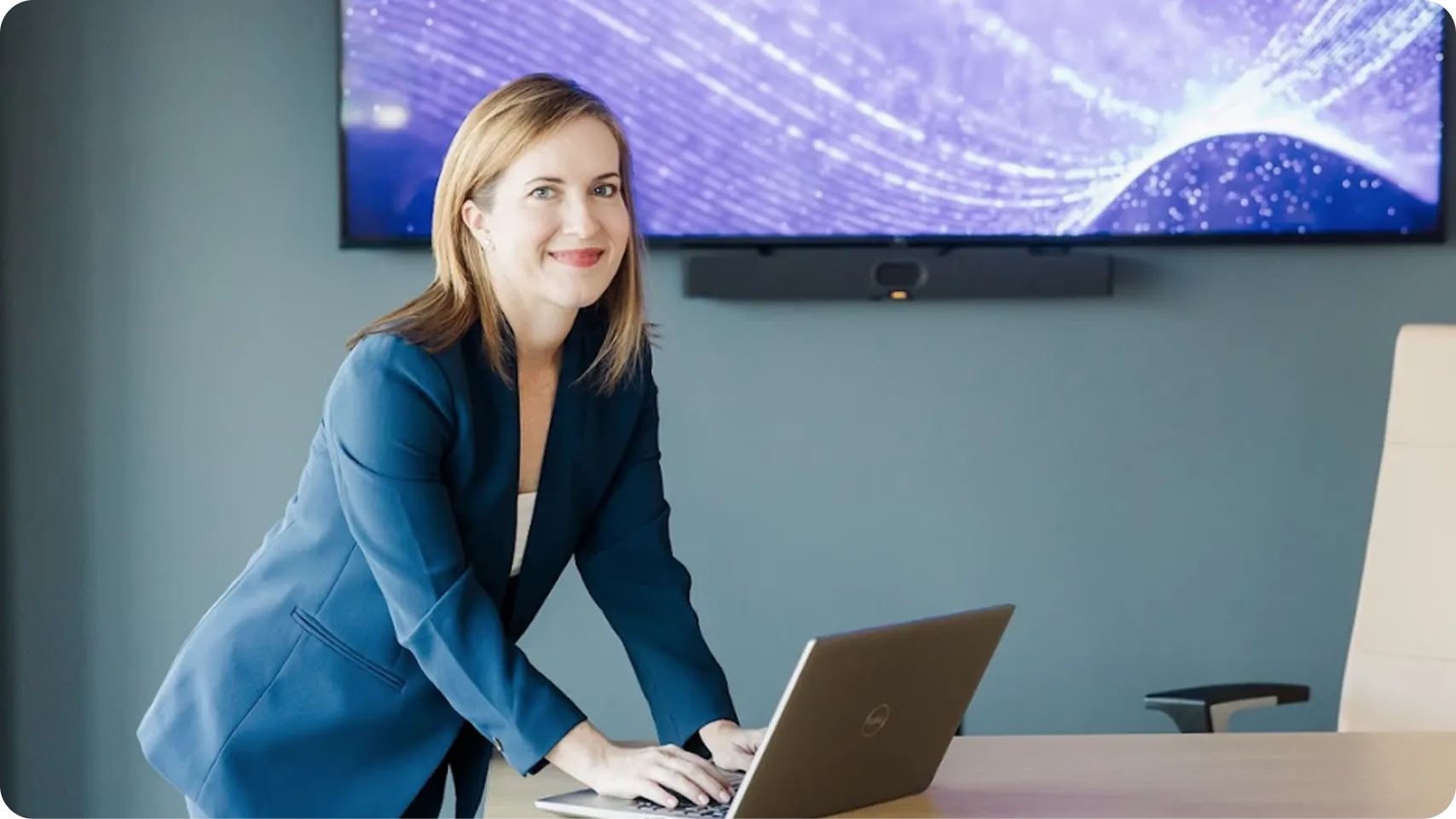 Smiling woman in blue blazer standing and typing on a laptop in a modern office with a large screen on the wall behind her.