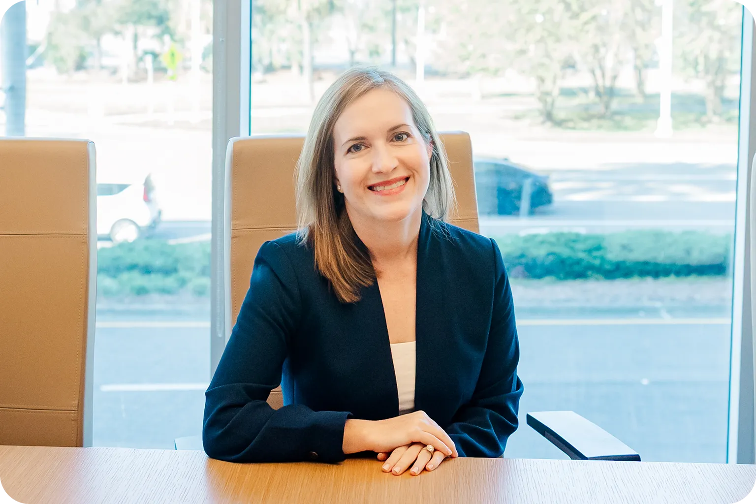 Smiling woman with light brown hair wearing a dark blazer sitting at a wooden table with a large window behind her.