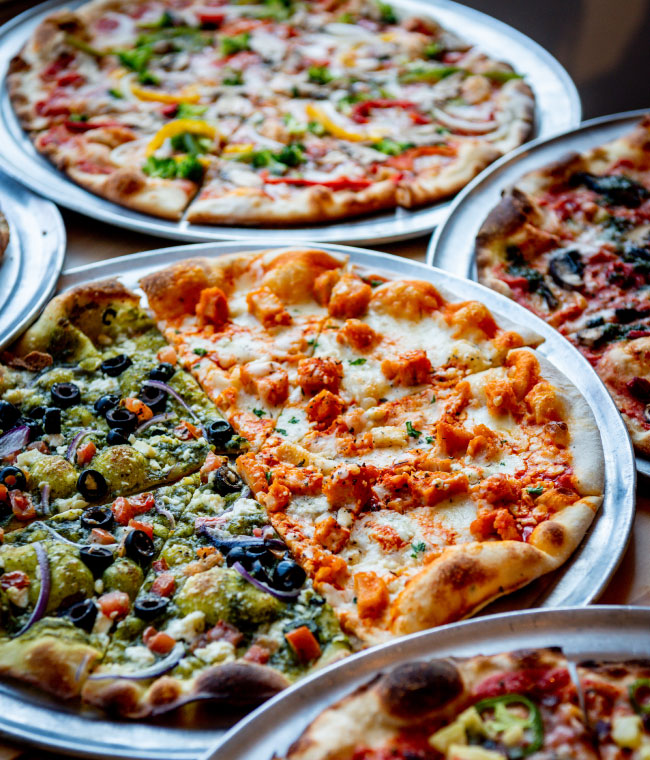 A variety of pizzas on metal trays, showcasing vibrant toppings. One
has olives and green sauce; another features orange chicken pieces.