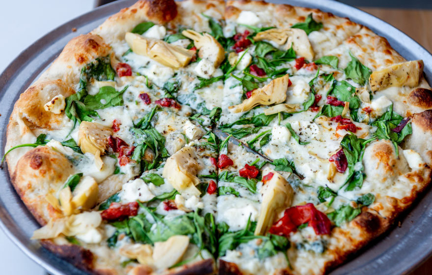 Delicious pizza topped with artichokes, fresh spinach, sun-dried tomatoes, and melted cheese on a thin crust, served on a metal tray.