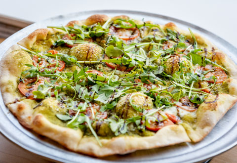 A freshly baked pizza topped with arugula, pesto, sliced tomatoes, and melted cheese on a crispy crust, presented on a metal tray.