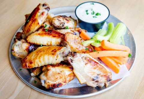 A plate of grilled chicken wings with crispy skin is served with carrot and celery sticks. A small cup of dipping sauce is on the side, garnished with herbs.