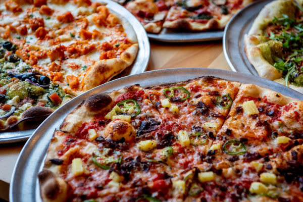 Array of assorted pizzas on silver trays, toppings include pepperoni, pineapple, jalapeños, veggies, and cheese.