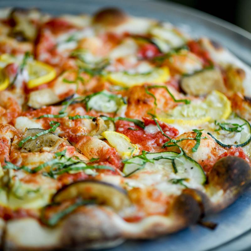 Close-up of a colorful pizza topped with sliced zucchini, yellow squash, fresh basil, and melted cheese on a crispy, golden-brown crust.