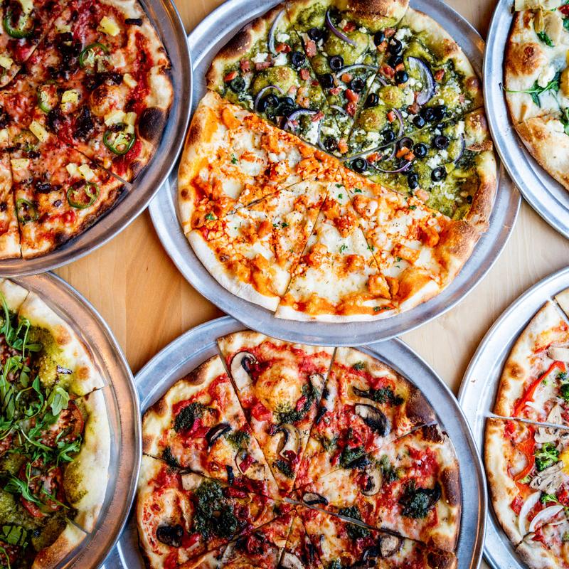 A variety of pizzas are arranged on wooden tables, each with unique toppings like olives, mushrooms, and arugula, creating a vivid and appetizing display.
