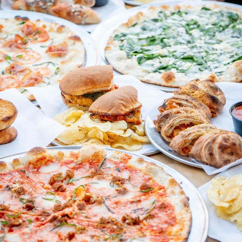 A variety of Italian-American foods on a table: pizzas with diverse toppings, a sandwich with chips, and a calzone with marinara sauce. A casual dining atmosphere.