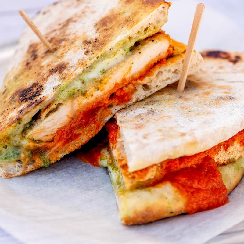 A close-up of a sliced panini sandwich on parchment paper. It features grilled chicken, vibrant red tomato sauce, and green pesto, secured with toothpicks.