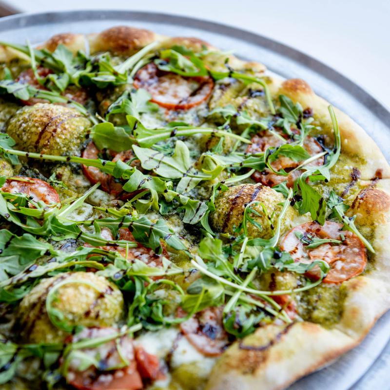 A fresh arugula and pesto pizza topped with tomato slices and mozzarella. Drizzled with balsamic glaze, it sits on a metal tray, exuding a rustic and vibrant appeal.