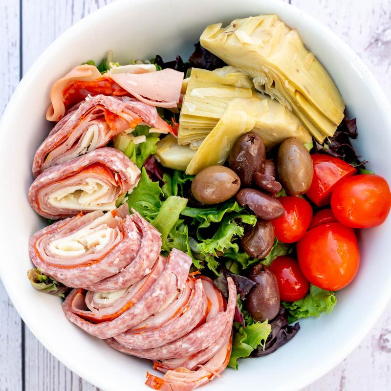 A vibrant salad in a white bowl with rolled meats, artichoke hearts, olives, cherry tomatoes, and mixed greens, creating a fresh, Mediterranean feel.