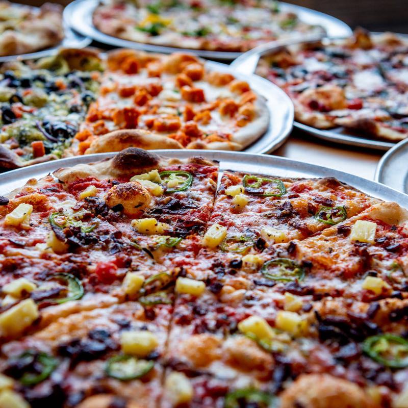 Close-up of various pizzas on metal trays, featuring toppings like pineapple, jalapeños, and pepperoni. The scene is vibrant and appetizing.
