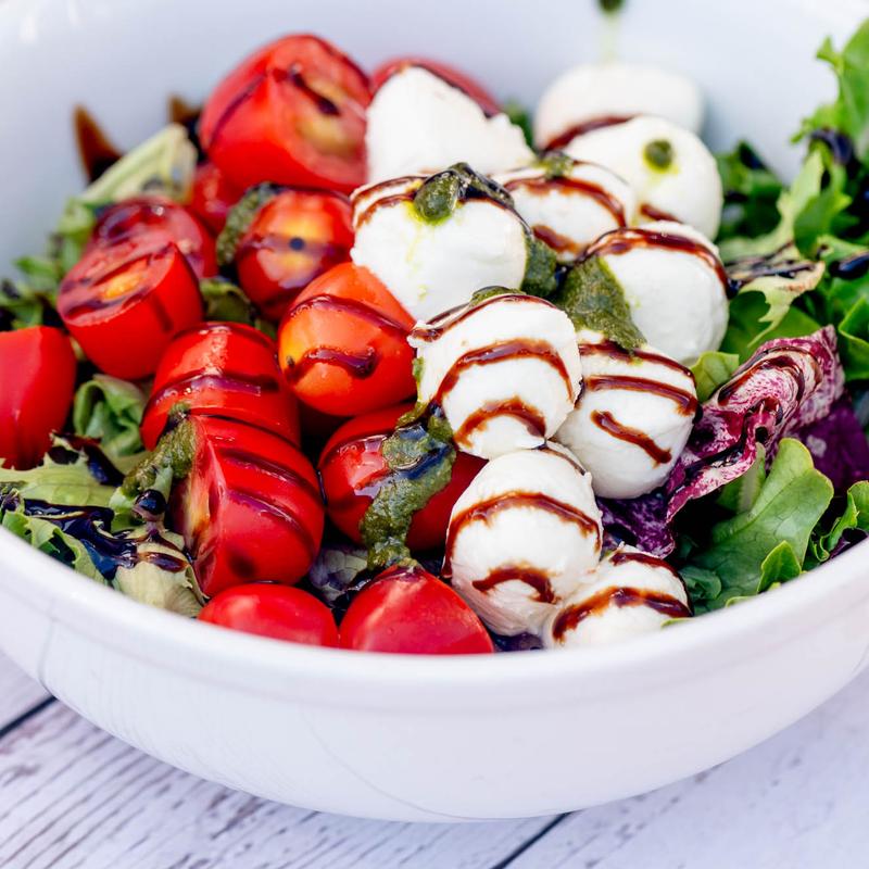 A fresh salad with cherry tomatoes, mozzarella balls drizzled with balsamic glaze, and pesto on leafy greens in a white bowl, creating a vibrant, appetizing look.