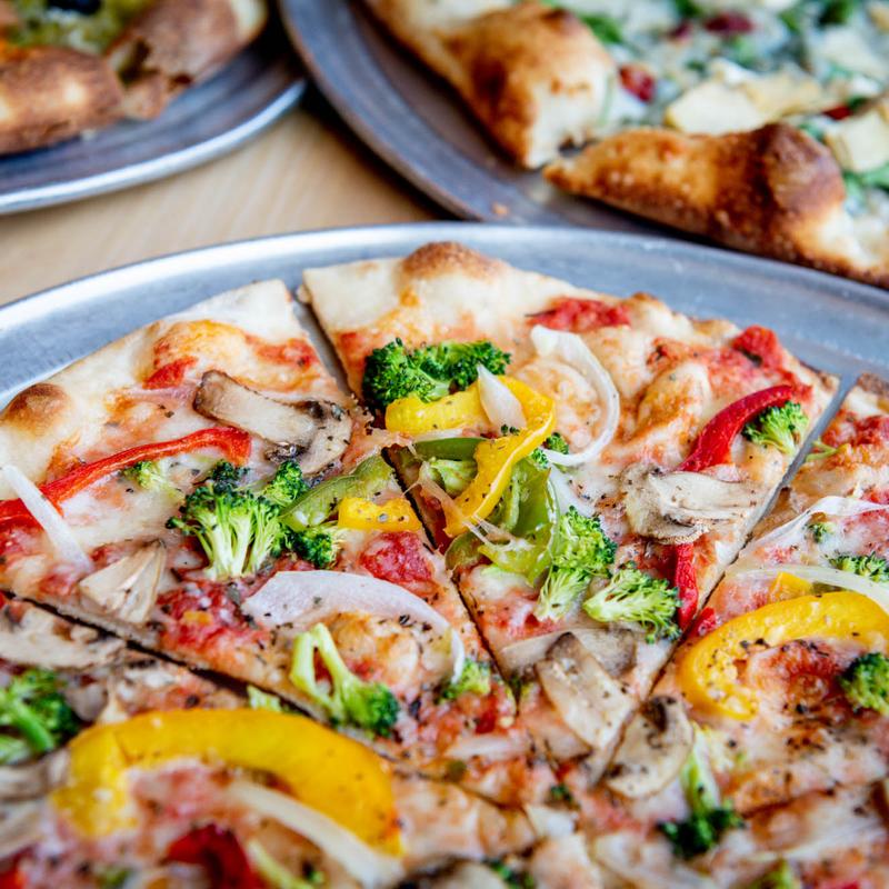 A vibrant veggie pizza topped with bright broccoli, yellow and red bell peppers, onions, and mushrooms on a thin crust. Background shows more sliced pizzas.