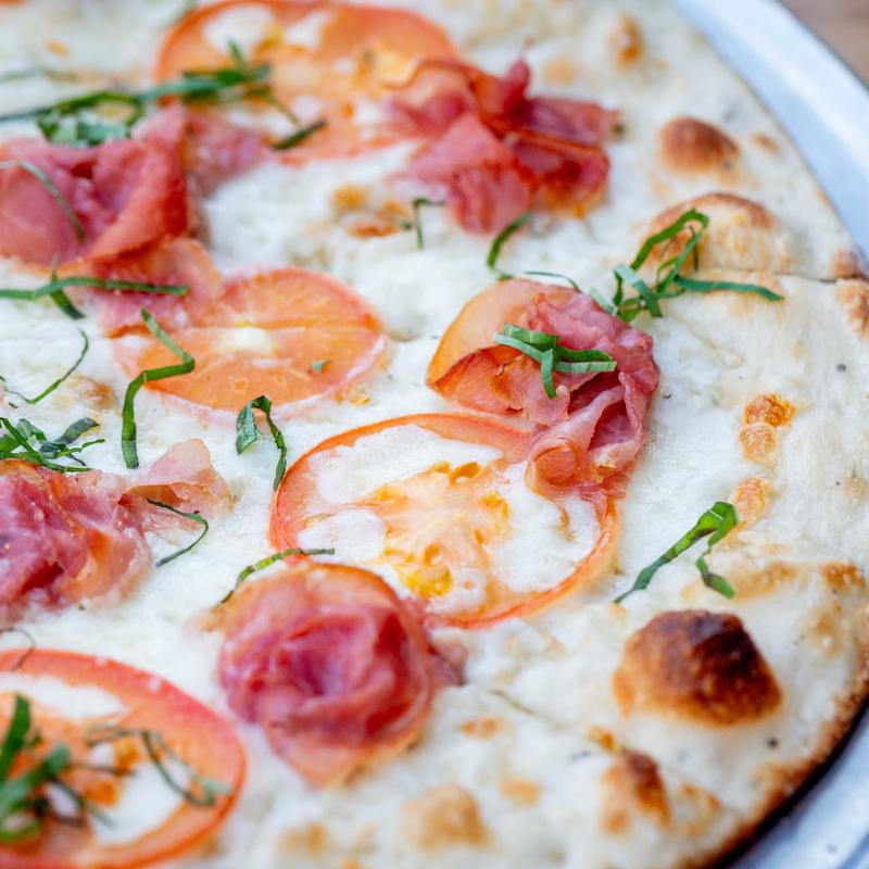 Close-up of a white sauce pizza with fresh tomato slices, prosciutto, and garnished with strips of basil. The crust is golden and slightly charred.