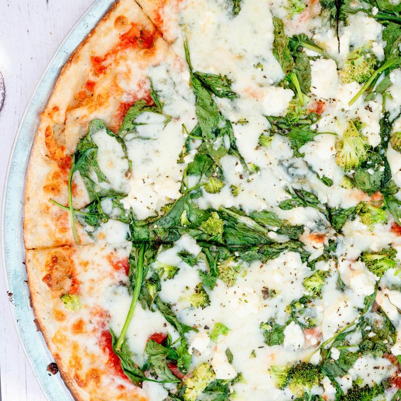 Close-up of a pizza topped with melted mozzarella, fresh spinach, broccoli, and tomato sauce. The vibrant green and red colors create an appetizing appearance.