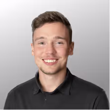 Junger Mann mit kurzem braunem Haar und schwarzem Hemd lächelt vor grauem Hintergrund.