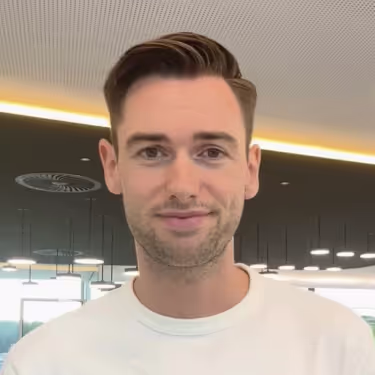 Porträt eines lächelnden jungen Mannes mit kurzen braunen Haaren in einem hellen Raum mit moderner Beleuchtung.