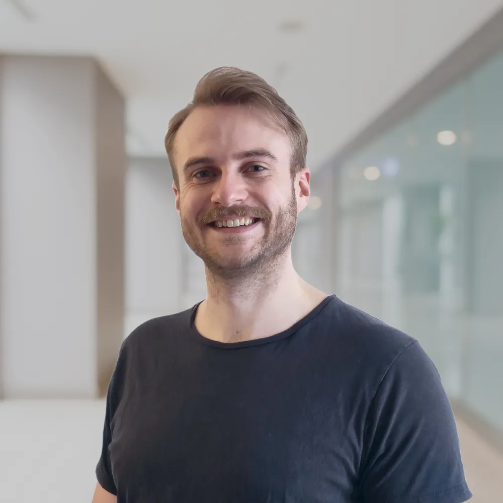 Lächelnder Mann mit kurzem braunem Haar und Bart in schwarzem T-Shirt vor unscharfem Bürohintergrund.