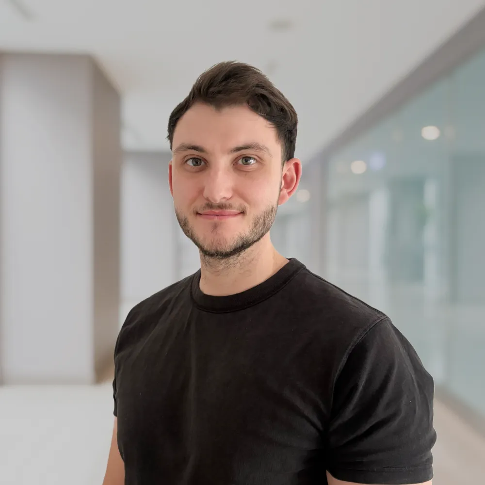 Porträt eines jungen Mannes mit kurzem dunklem Haar und Bart, der ein schwarzes T-Shirt trägt und in einem modernen, hellen Innenraum steht.
