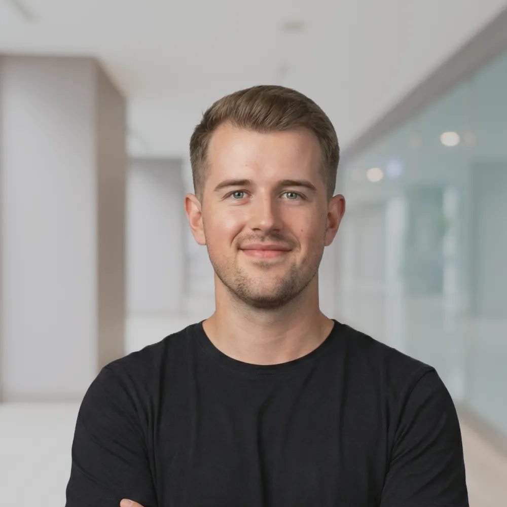 Lächelnder junger Mann mit kurzen braunen Haaren und schwarzem T-Shirt vor einem modernen Bürohintergrund.