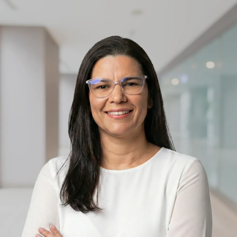 Lächelnde Frau mit langen dunklen Haaren, klaren Brillen und weißem Oberteil vor einem modernen Bürohintergrund.