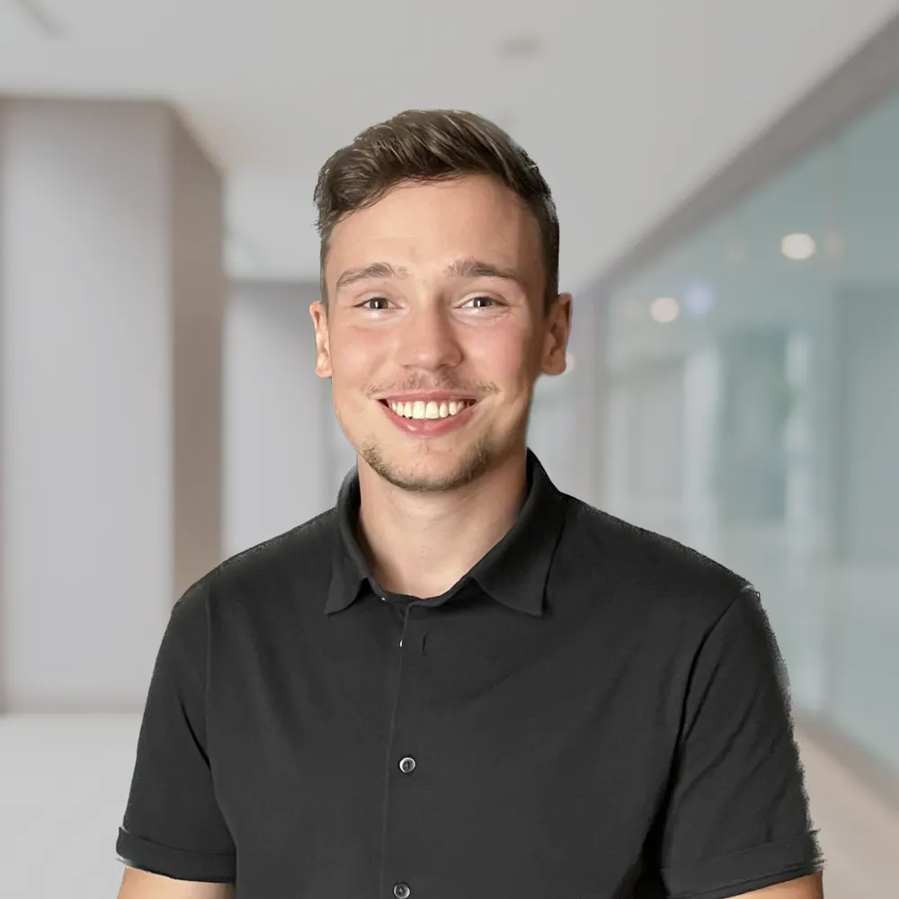 Junger Mann mit kurzen braunen Haaren und schwarzem Hemd lächelt in einem modernen Büroflur.