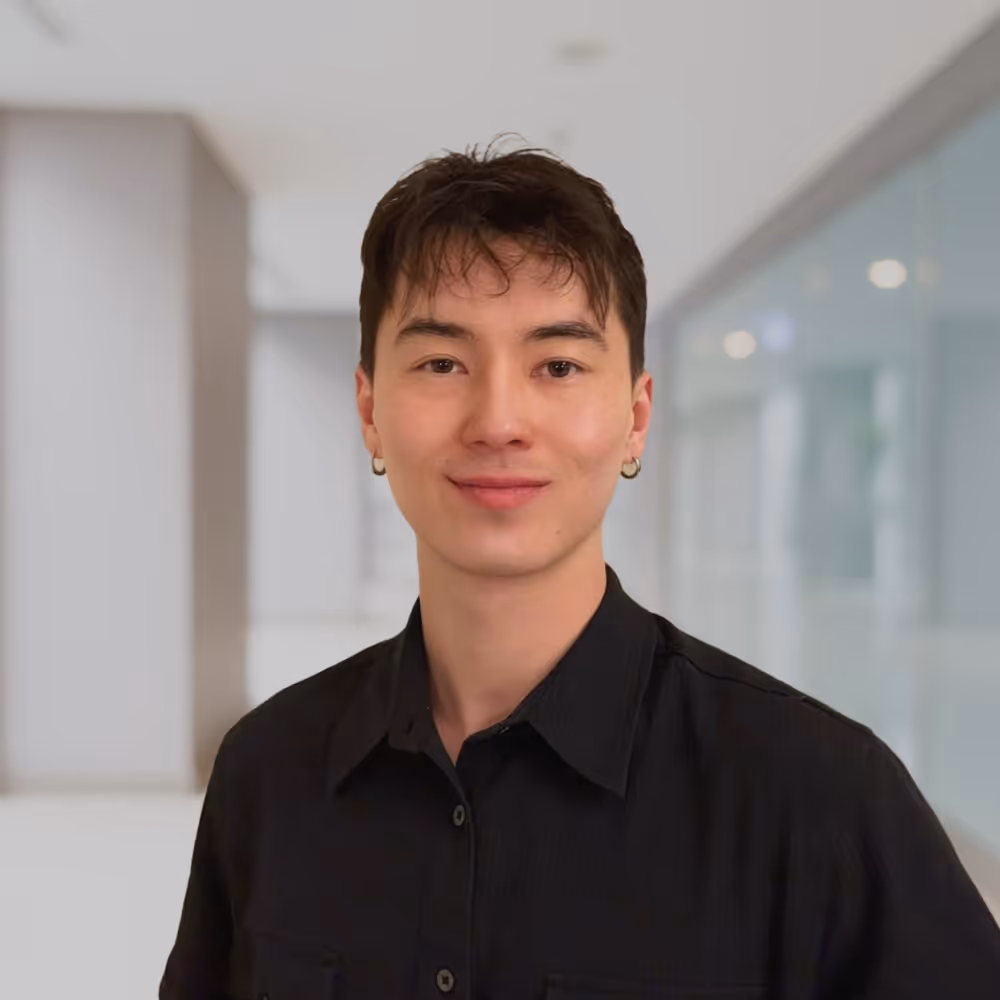 Porträt eines jungen Mannes mit kurzen dunklen Haaren, der ein schwarzes Hemd trägt und in einem modernen Bürogebäude lächelt.