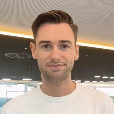 Junger Mann mit kurzem braunem Haar und schwarzem Hemd lächelt vor grauem Hintergrund.