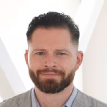 Porträt eines jungen Mannes mit dunklen Haaren, der ein schwarzes Shirt vor einem grauen Hintergrund trägt.