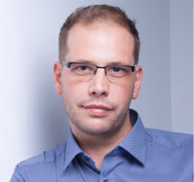 Junger Mann mit kurzem braunem Haar und schwarzem Hemd lächelt vor grauem Hintergrund.