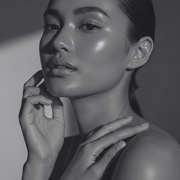 Black and white close-up portrait of a woman with glowing skin, posed with one hand touching her face and the other on her shoulder.