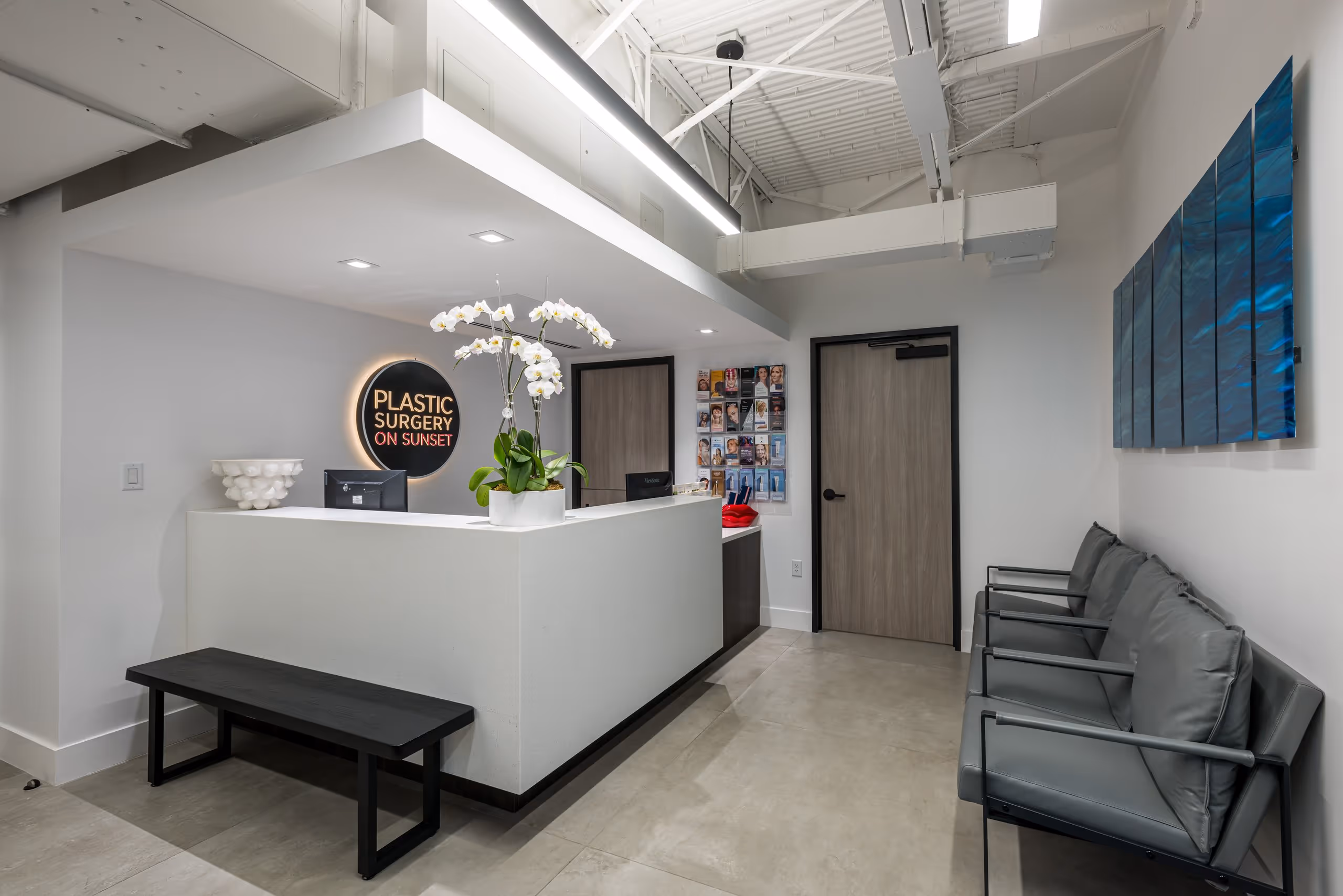 Modern clinic reception with white desk, orchid plant, black bench, gray chairs, and Plastic Surgery on Sunset sign.
