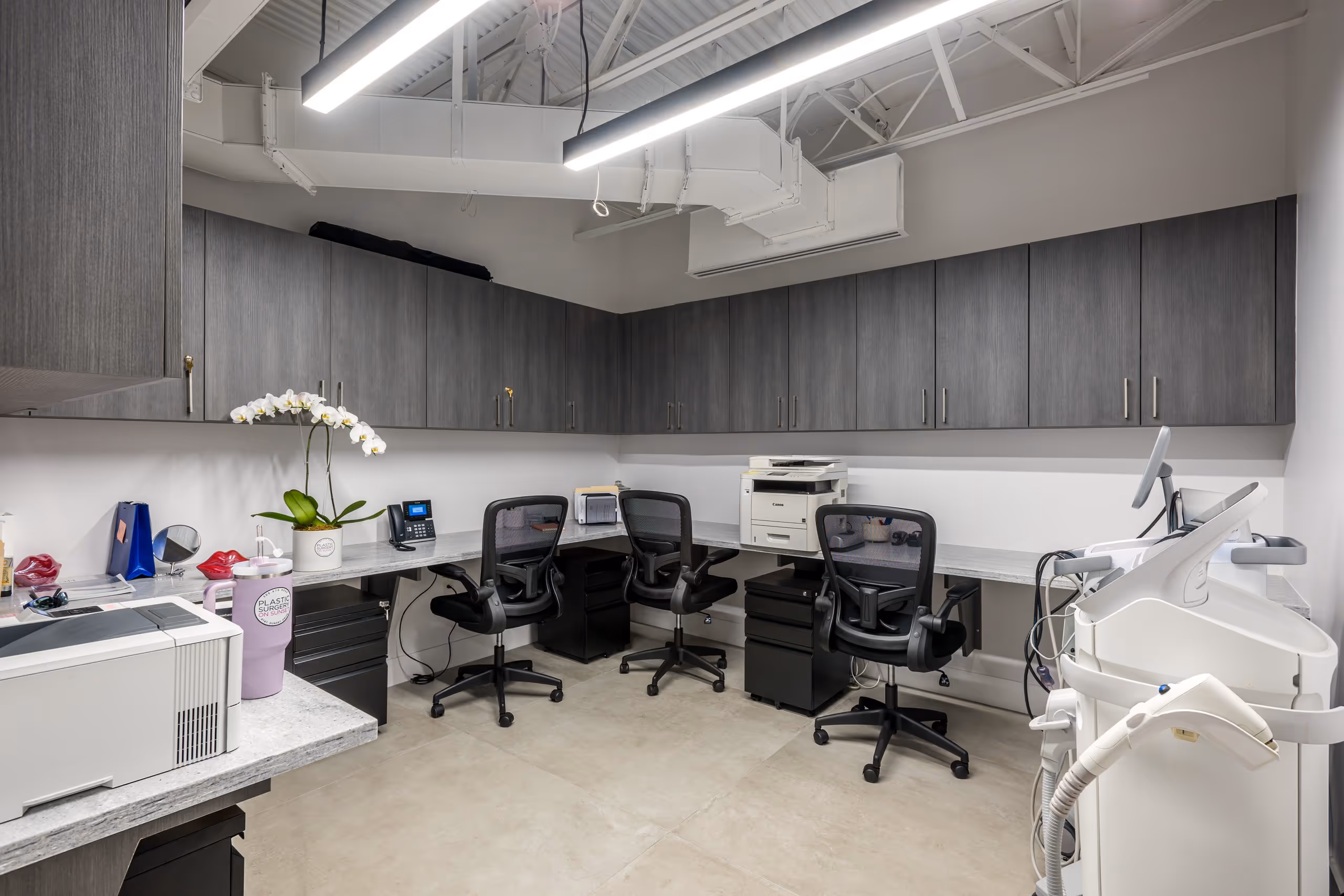 Modern office workspace with gray cabinets, three black swivel chairs, printers, and medical equipment under bright ceiling lights.