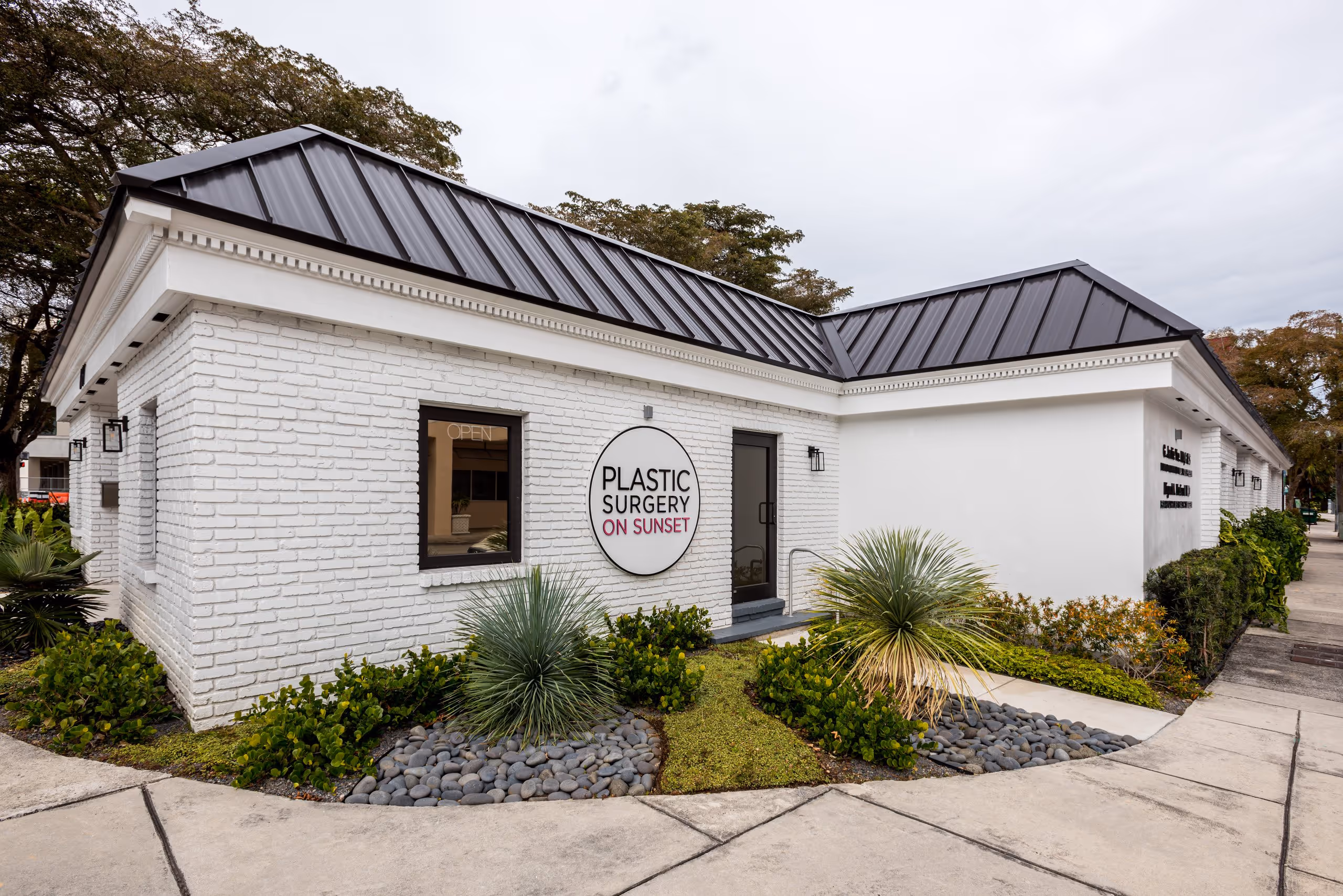 White brick building with black metal roof, sign reading 'Plastic Surgery on Sunset,' surrounded by landscaped greenery and rocks.