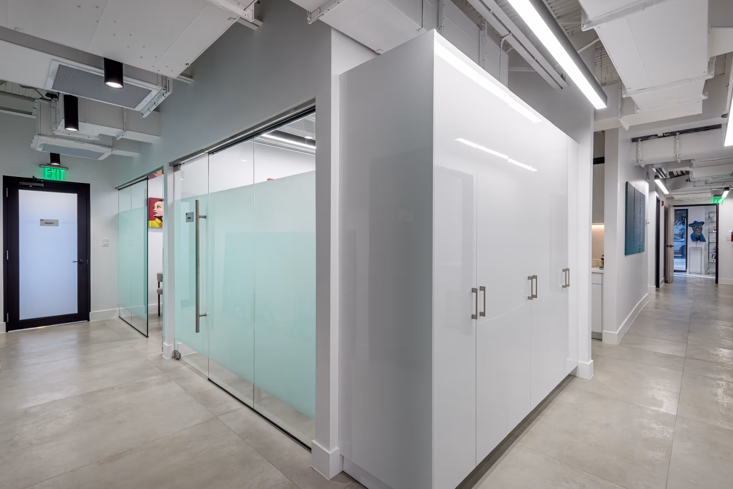 Modern office hallway with glass doors, white cabinets, gray floor tiles, and ceiling lights.