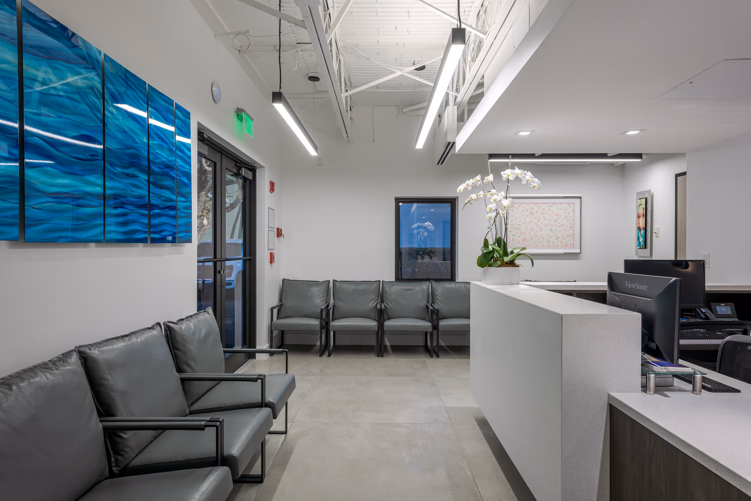 Modern waiting room with gray chairs, white reception desk with orchid, and blue abstract wall art.