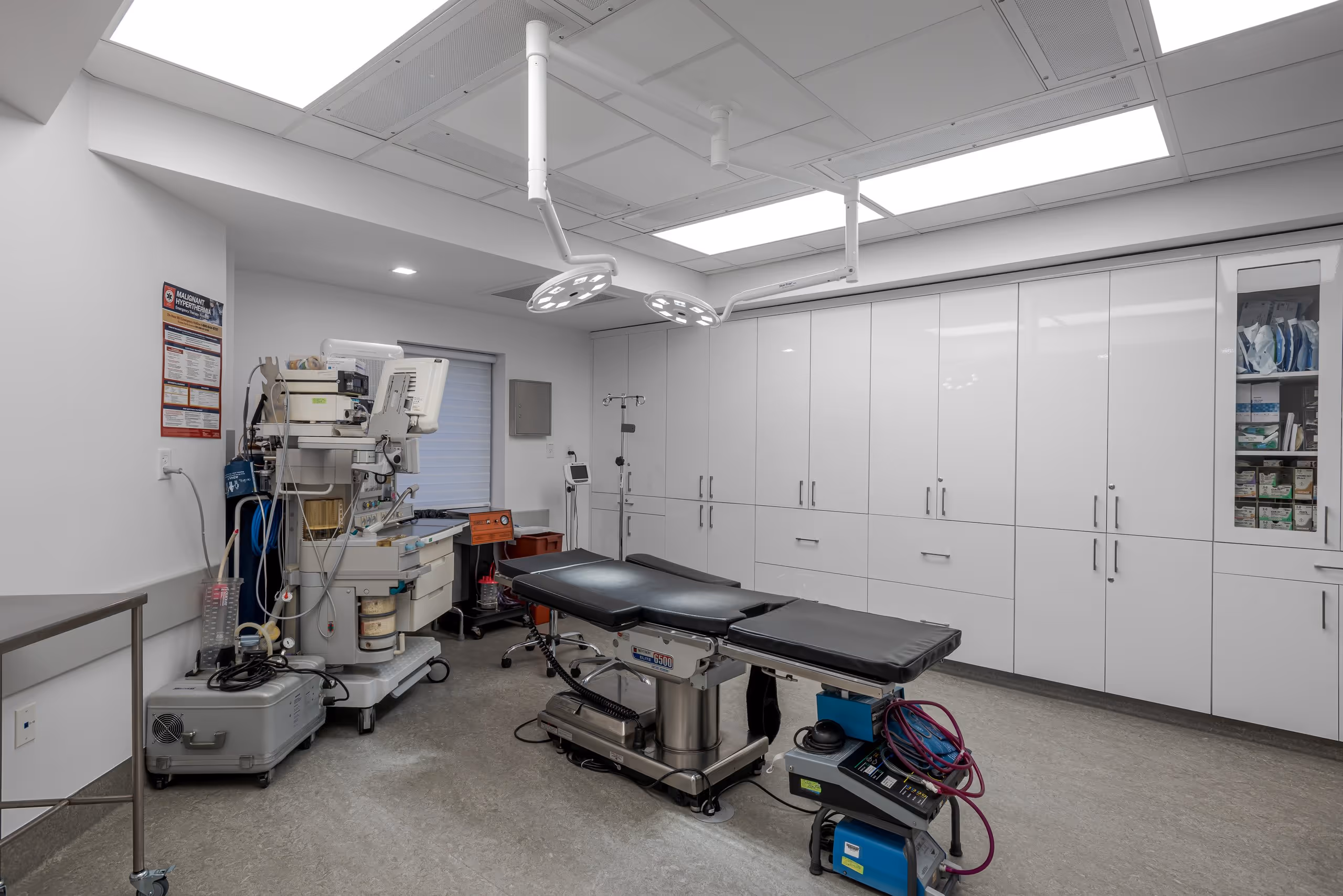 Modern operating room with surgical table, overhead surgical lights, medical equipment, and white storage cabinets.