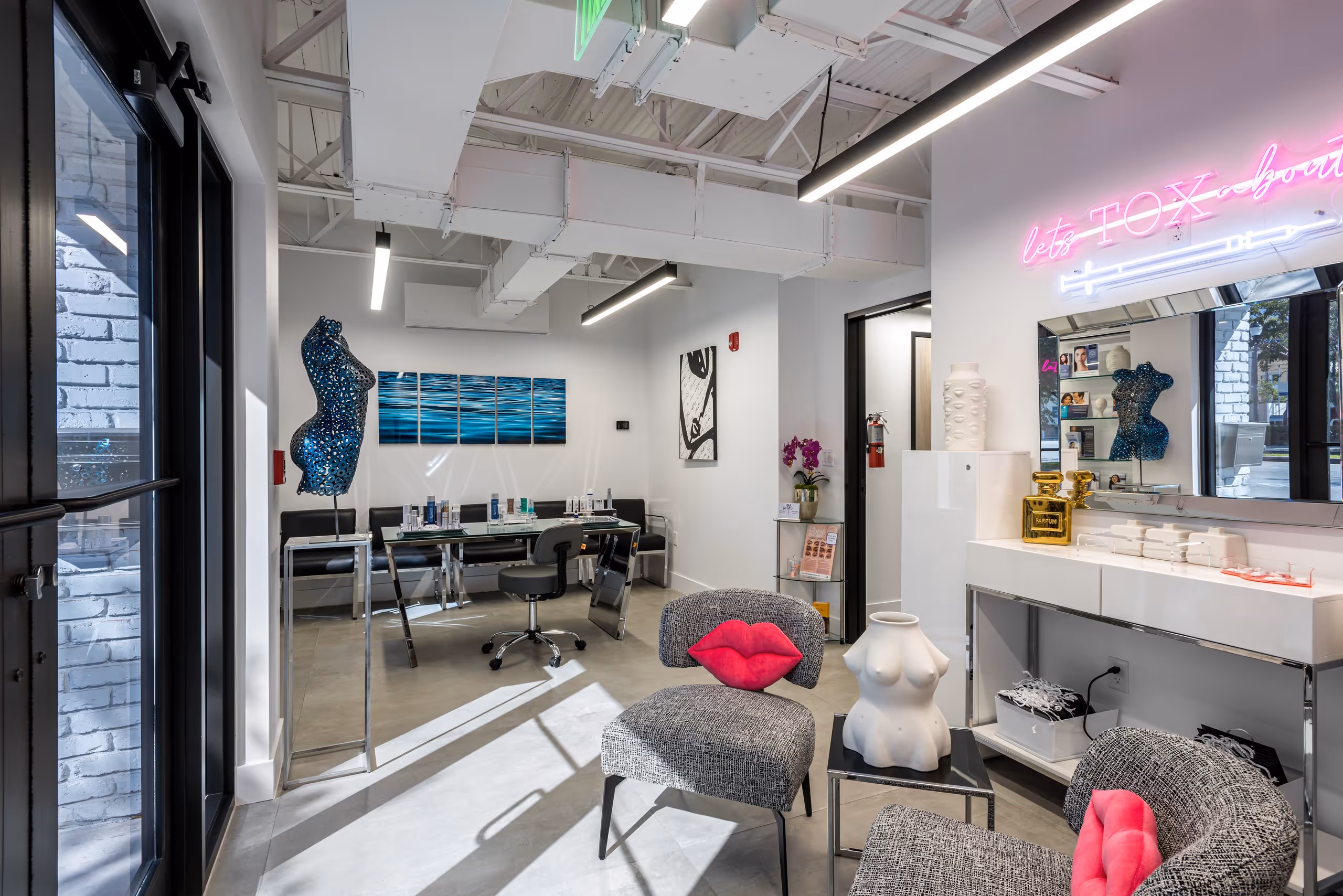 Modern cosmetic or beauty clinic interior with gray chairs featuring pink lip-shaped pillows, sculptural torso art pieces, and a neon sign reading 'let's TOX'.
